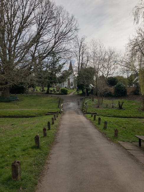 A paved pathway leading through a park-like area with grass and leafless trees on either side, bordered by small wooden posts and a few benches. In the background, there is a white building with a tall, pointed steeple, suggesting a church or similar structure. The sky is overcast, and the overall scene is calm and quiet. This setting is part of a house removal or relocation process facilitated by Man and Van Belsize Park, who are handling furniture transport and packing as part of the move from Englands Lane to Belsize Village in Belsize Park.