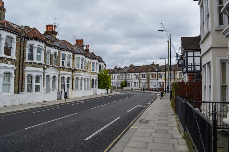 A quiet residential street with a row of Victorian-style terraced houses featuring white facades, bay windows, and tiled roofs, on the left side of the image. The pavement runs along the front of these properties, with a black wrought iron fence bordering one of the front gardens. On the right side, there is a modern white building with a black timbered extension and a small garden area. The street is lined with grey street lamps, and several parked cars are visible further down the road. The sky is overcast with grey clouds, casting diffuse lighting across the scene. In the background, a construction crane is visible above the rooftops, indicating ongoing development. The scene depicts an environment suitable for house removals and furniture transport, with no visible moving activity or equipment, but it reflects the typical setting for home relocation activities conducted by companies like Man and Van Belsize Park, especially involving the process of packing, loading, or transporting furniture and boxes from properties situated along this street.