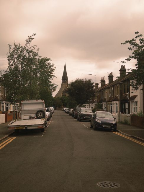 A paved pathway leading through a park-like area with grass and leafless trees on either side, bordered by small wooden posts and a few benches. In the background, there is a white building with a tall, pointed steeple, suggesting a church or similar structure. The sky is overcast, and the overall scene is calm and quiet. This setting is part of a house removal or relocation process facilitated by Man and Van Belsize Park, who are handling furniture transport and packing as part of the move from Englands Lane to Belsize Village in Belsize Park.